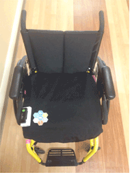 The image shows a manual wheelchair with a pressure mat draped over the seat cushion. The map is made of a water resistant, 4-way stretch material that covers the seat cushion entirely and fits snug with use of elastic around the bottom edge.  There is a USB cable extending from the pressure map, leading to a small box that is approximately 2"x5"x1".  The box contains the battery power and the mini-PC.  